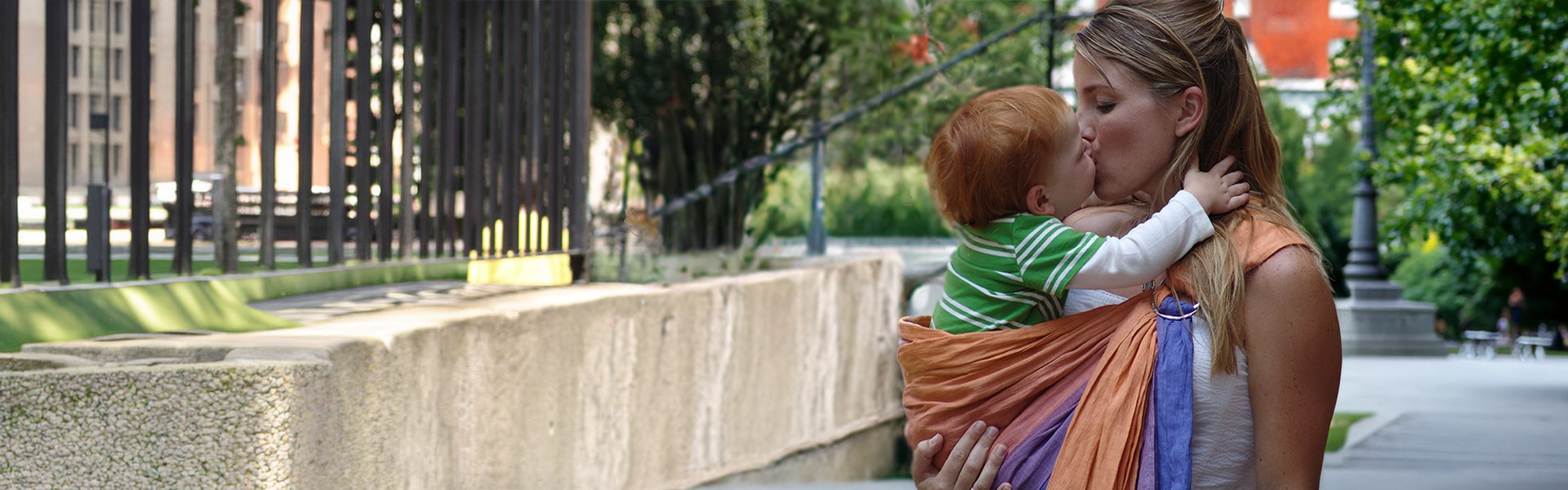 Mother kissing her child while carrying them in a ring sling, expressing closeness and lasting memory